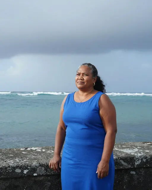 Middle aged Pacific Islander woman in a blue dress stands on a harbor wall as storm clouds clear over the ocean