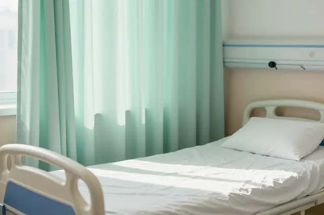Morning sunlight falls across a made hospital bed next to a half-drawn pale green privacy curtain