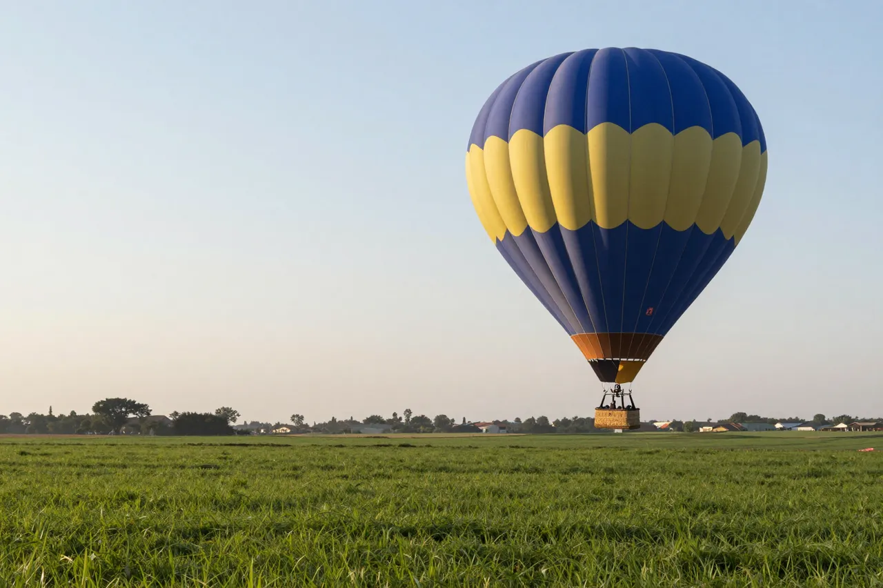 Blue and yellow hot air balloon ascending over a green grassy field at pale dawn