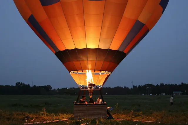 A hot air balloon glows from its burner as it inflates on a grassy meadow at pre-dawn twilight