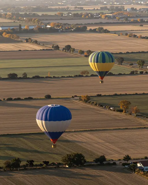 Two hot air balloons drift low over patchwork autumn farm fields during golden sunrise