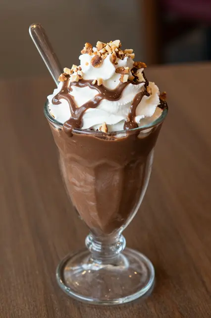 A tall glass hot fudge sundae topped with whipped cream, chocolate sauce, and chopped nuts sits on a wooden table.