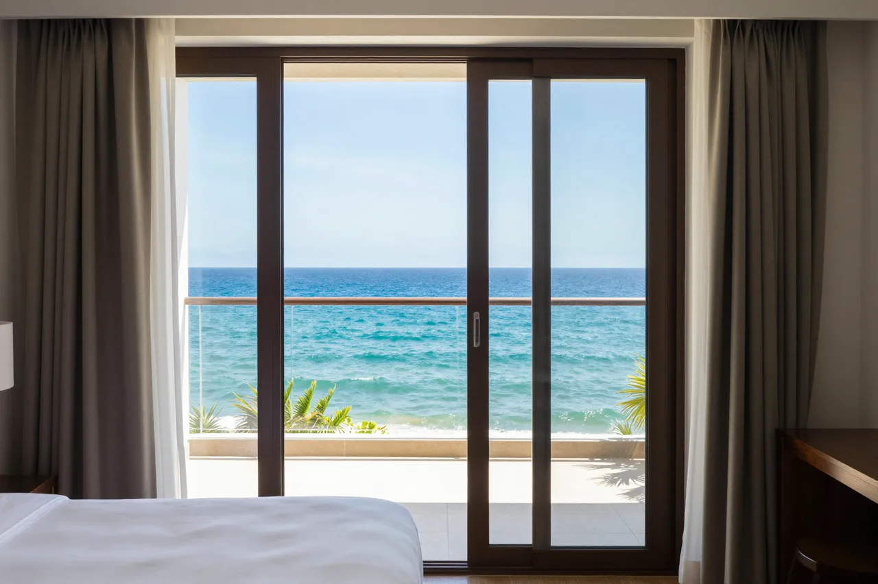 A modern hotel bedroom looks out through sliding glass doors to a sunny turquoise ocean view