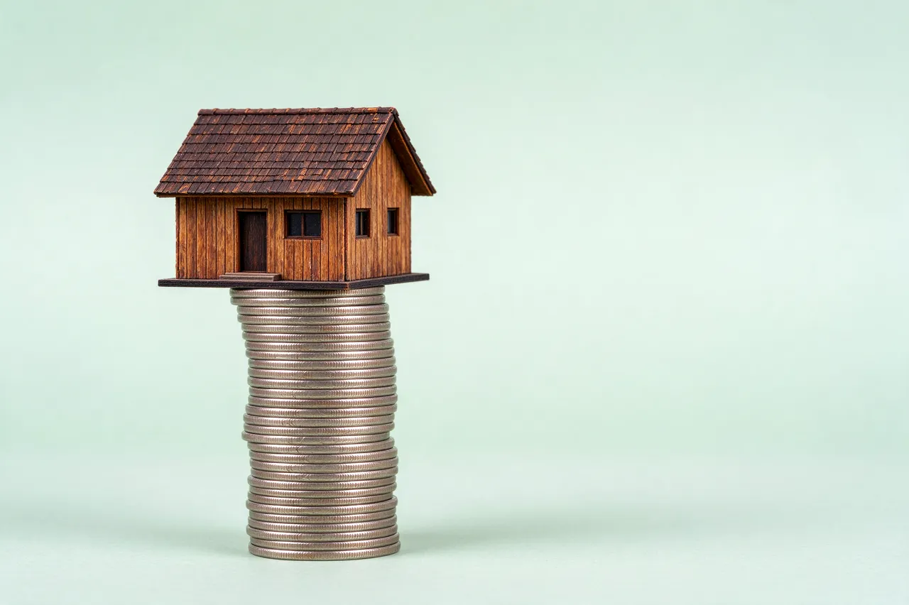 A small wooden house model rests on top of a tall stack of coins against a pale green background.