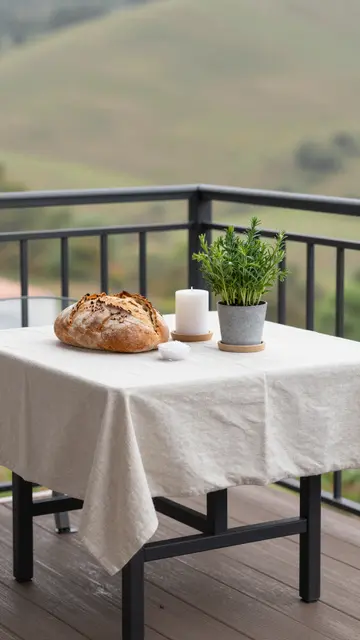 A linen-covered balcony table holds sourdough bread, salt, a candle and potted herb with rolling hills beyond
