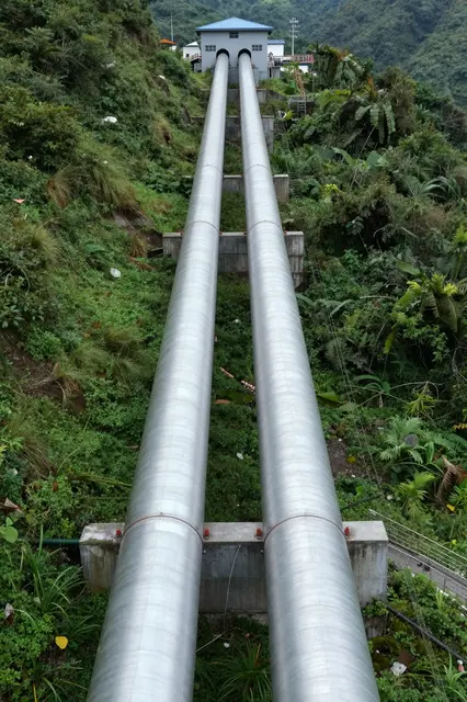 Two large steel water pipes run down a steep vegetated slope to a small hydroelectric power station.