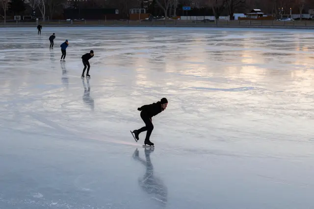 Group of speed skaters gliding across a smooth frozen pond at golden sunset, seen from a distance.