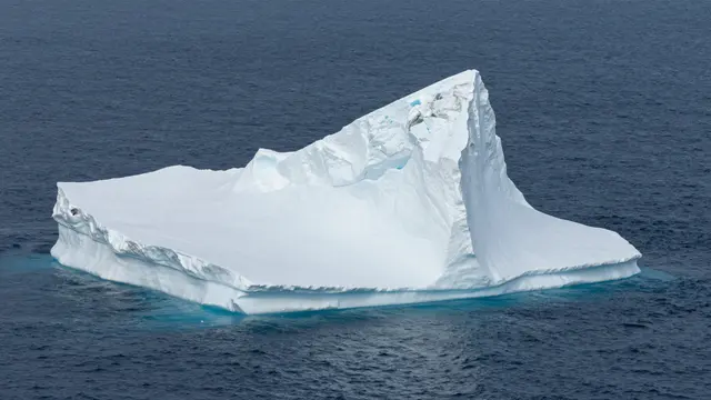 A large angular white iceberg with faint blue crevices floats in dark deep blue arctic sea water.