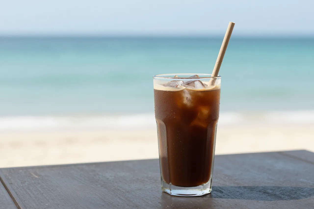 A clear glass of iced coffee with a paper straw sits on a wooden beach table overlooking blurred turquoise sea.