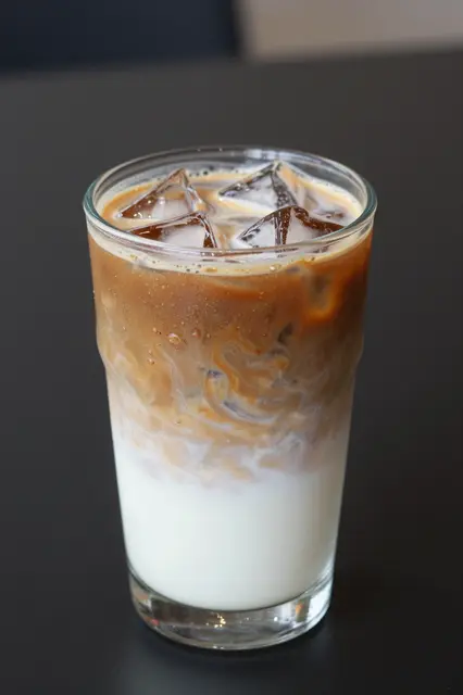 A tall clear glass of iced coffee with a swirling milk layer sits on a dark table