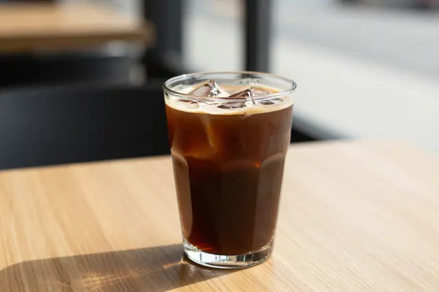 A clear faceted glass of iced cold brew coffee sits on a sunlit wooden cafe table.