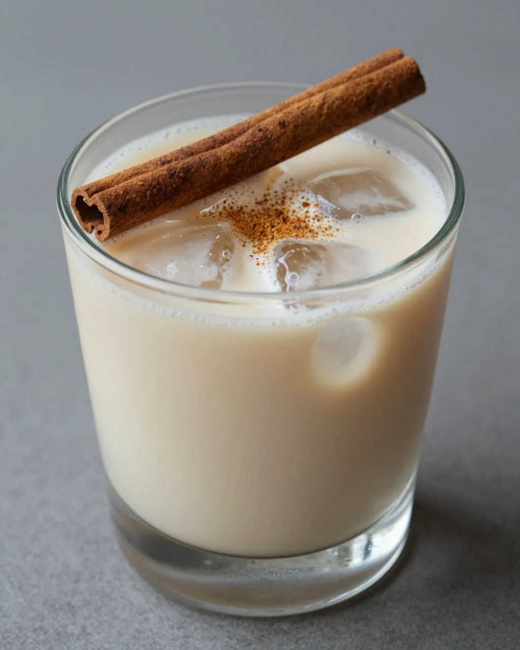 A clear glass of iced creamy horchata topped with cinnamon sticks and ground spice on a gray surface