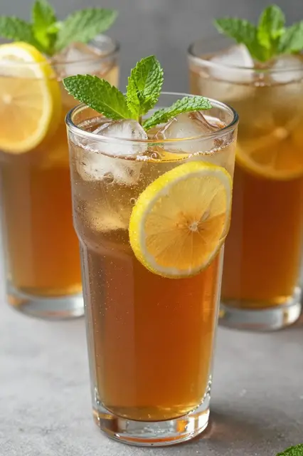 Three tall glasses of iced tea garnished with lemon slices, mint, and ice sit on a gray countertop.