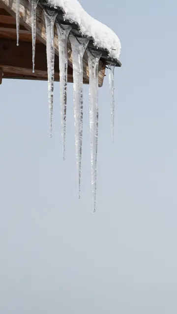 Clear icicles hang from a snow-dusted wooden roof eave against a pale overcast winter sky with empty copy space below.