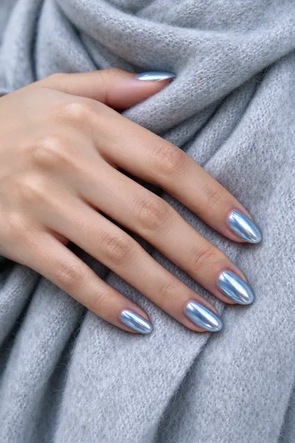 A hand with pale olive skin resting on soft pale grey wool fabric, showing shiny almond icy blue chrome nails.