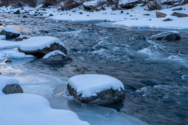 A fast flowing icy river passes snow covered rocks and thin ice at cold blue hour