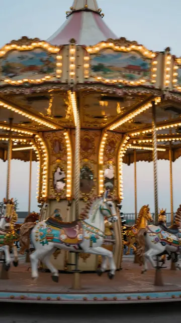 Ornate lit carousel with painted horses stands under a pale sky at twilight
