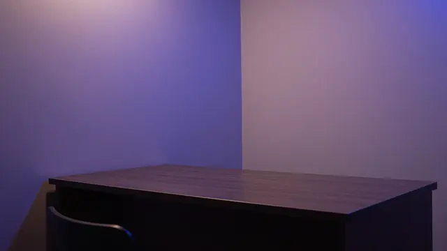 An empty wooden desk sits in a dark room corner lit with soft violet and blue ambient wall light