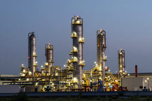 Lit oil refinery distillation towers stand against a pale blue evening sky, viewed from a distance
