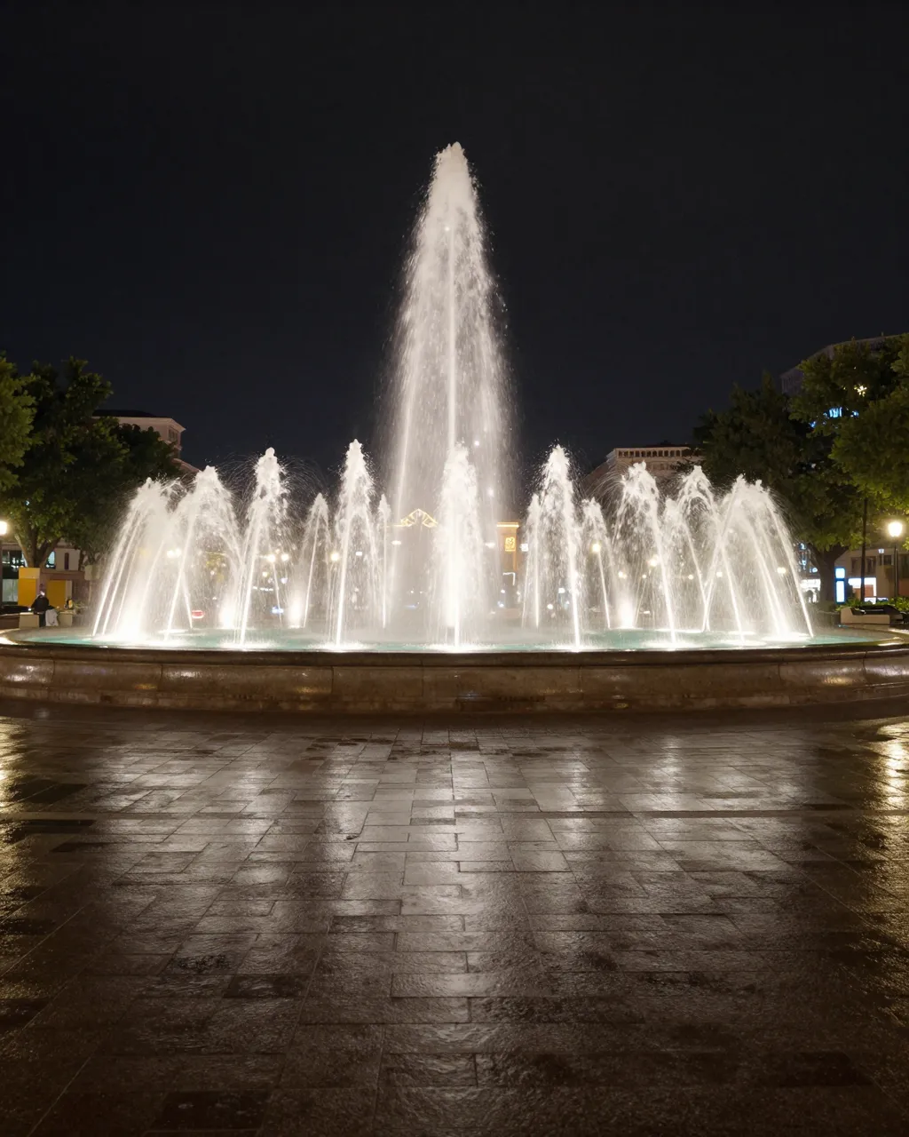 A lit multi-jet fountain sits on wet reflective paving in an empty night city plaza