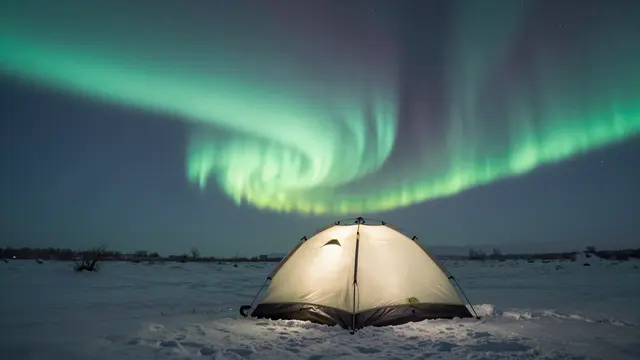 Lit camping tent sits on a snowy plateau beneath rippling green aurora and starry night sky