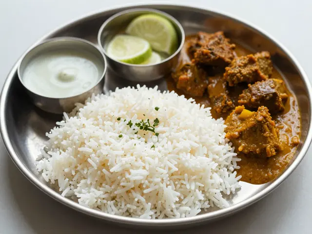A traditional stainless steel Indian thali holds basmati rice, meat curry, raita, and lime wedges on a light table surface.