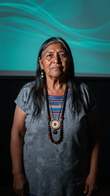 Indigenous woman in her sixties stands in a dark room with a teal projected light backdrop