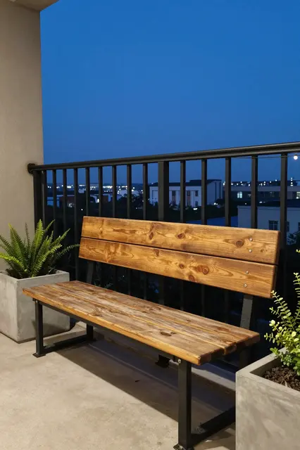 Reclaimed wood bench sits on a concrete balcony next to planters under a black steel railing at twilight