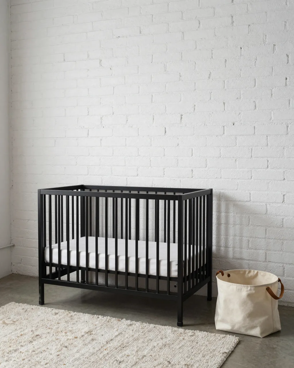 Black metal crib sits next to a canvas storage basket and wool rug against a white painted brick wall