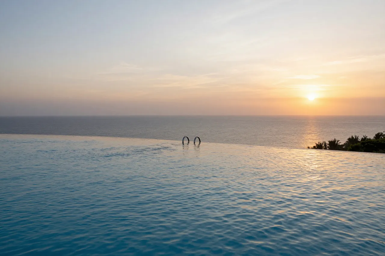 Rippled infinity pool overlooks a calm ocean under a warm setting sun, with distant palm trees on the horizon