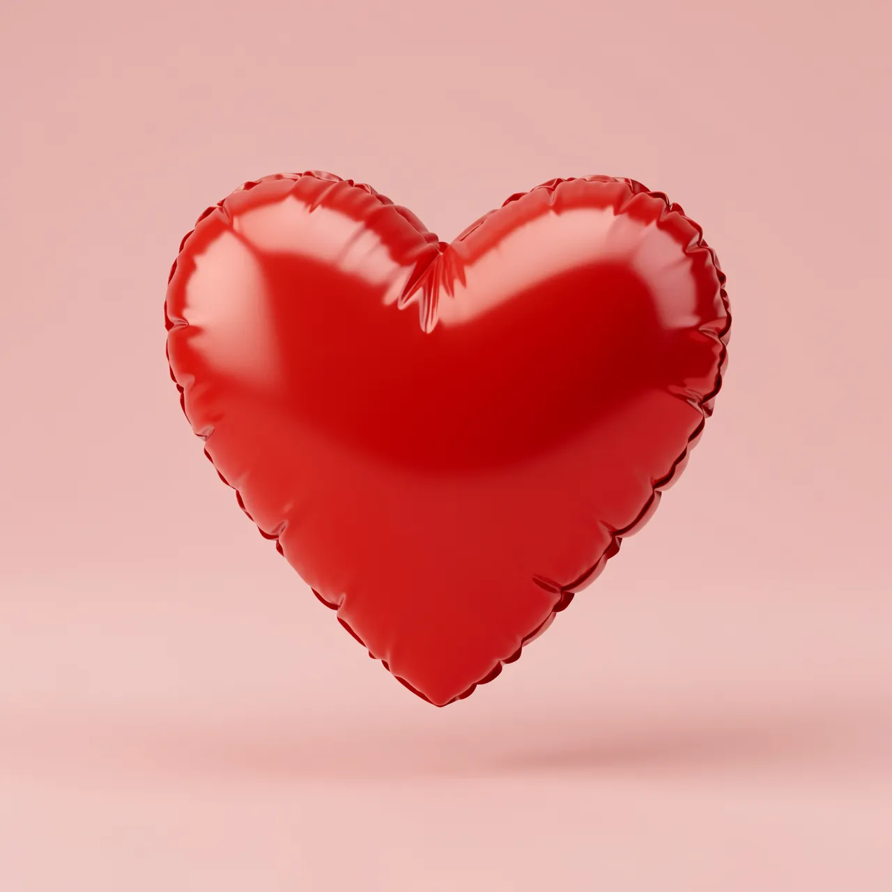 Glossy puffy red inflatable heart floats centered against a soft blush pink studio background