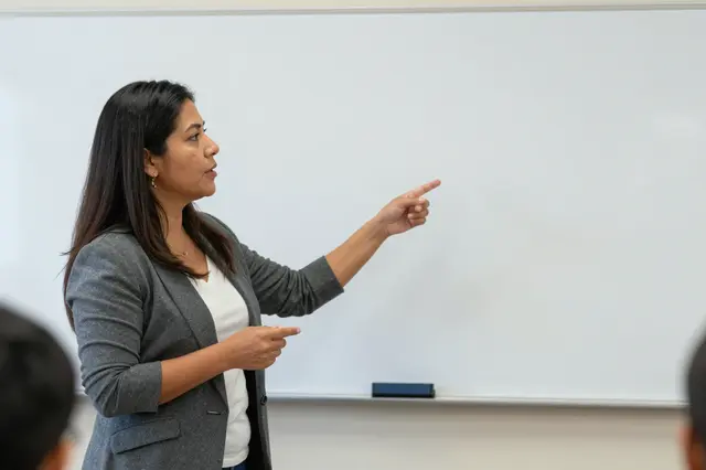 Side view of a Latina woman in her forties gesturing while lecturing next to a blank whiteboard in a community room.