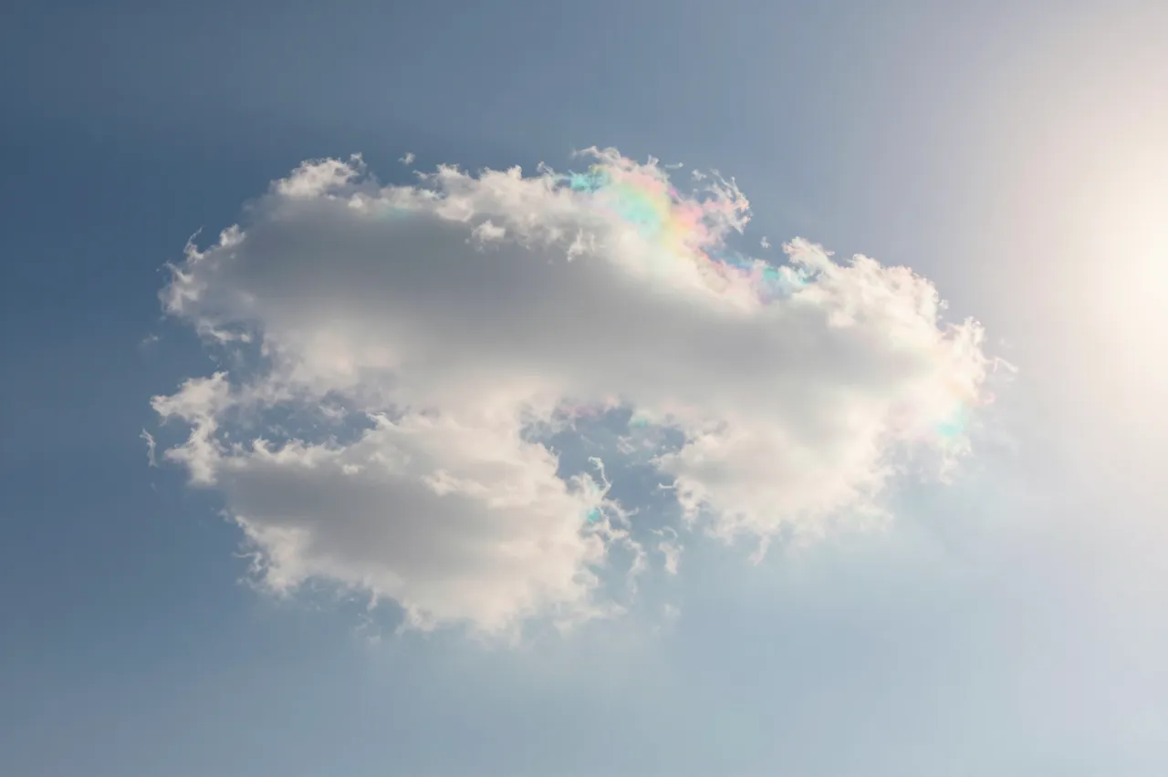 A fluffy white cloud displays iridescent pastel shimmer next to bright sunlight in a pale blue sky