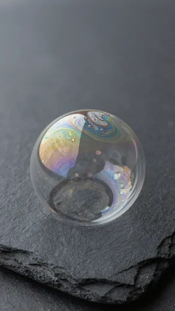 A single spherical soap bubble with swirling rainbow iridescence rests on a dark textured slate surface