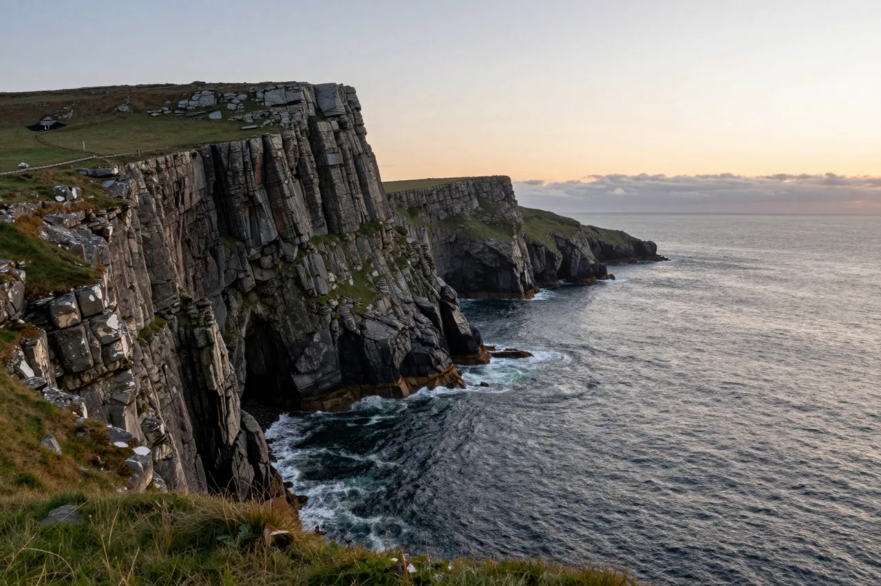 Rugbed layered sea cliffs rise above the Atlantic Ocean under a soft sunset sky