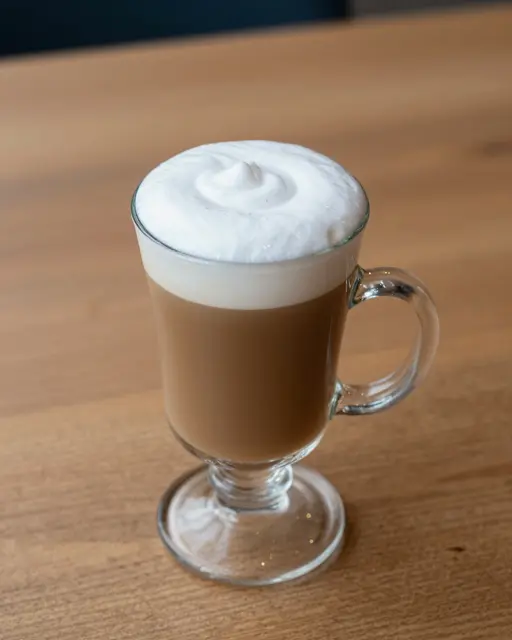 A glass mug of Irish coffee topped with thick swirled cream sits on a warm wooden table