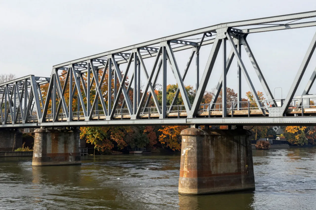 Gray steel truss bridge spans a calm river with colorful autumn trees in the background under a pale sky