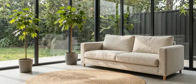 Bright japandi sunroom with linen sofa, jute rug and potted maple saplings next to floor-to-ceiling glass