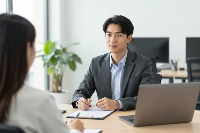 A hiring manager sits across a table from a back-facing candidate during an office job interview