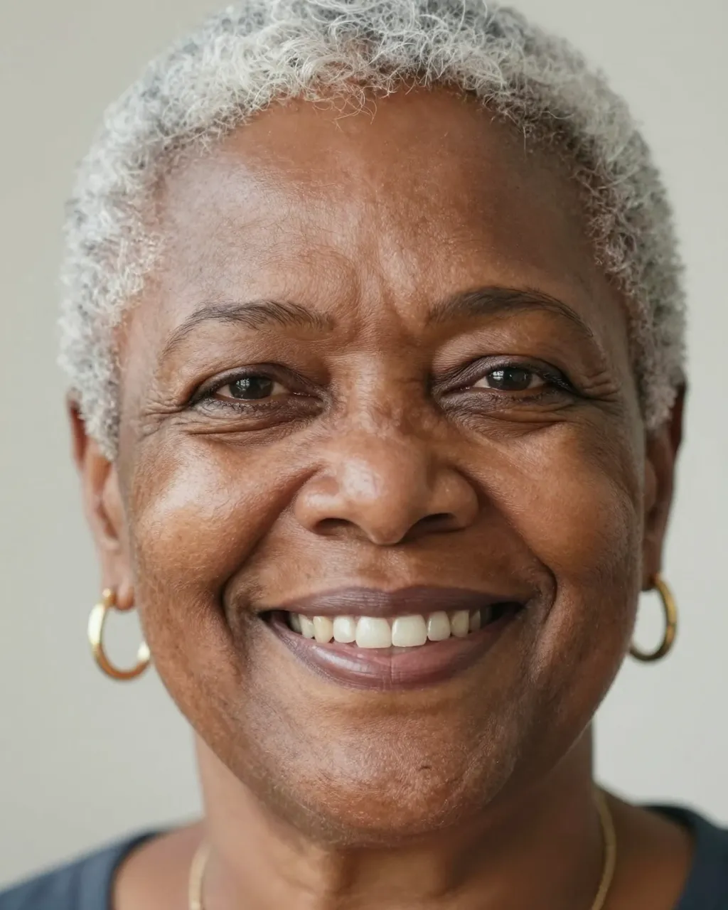 Close-up portrait of a smiling senior Black woman with short silver hair and gold hoop earrings under soft light