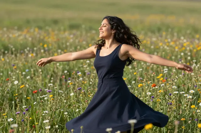 A joyful woman in a navy dress spins with arms outstretched in a sunlit wildflower meadow