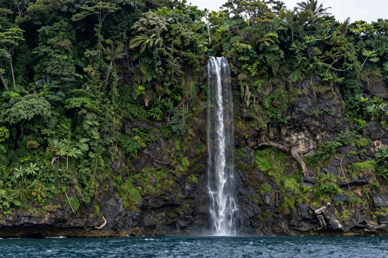 A tall waterfall cascades down a rocky jungle-covered cliff directly into dark blue ocean water.
