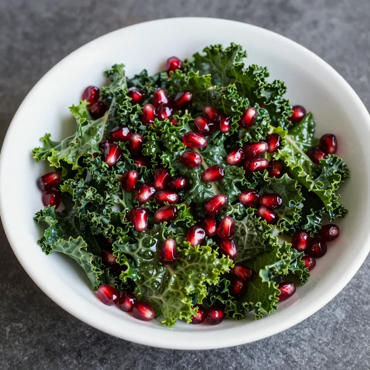 High angle view of a white bowl of fresh kale and pomegranate arils on a dark stone surface