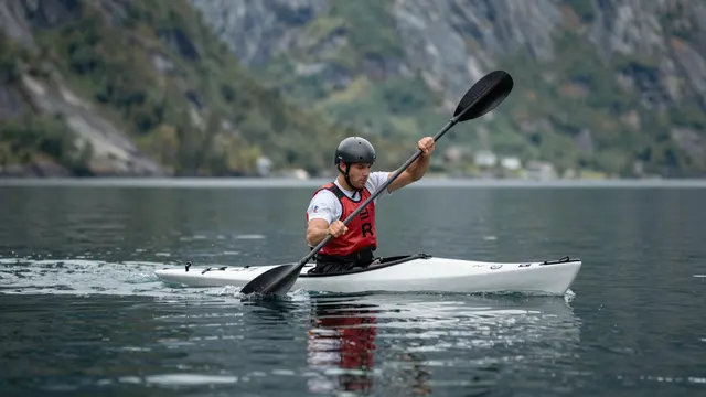 A male kayaker in a helmet and life vest paddles a white kayak across still fjord water with steep rocky mountains in the