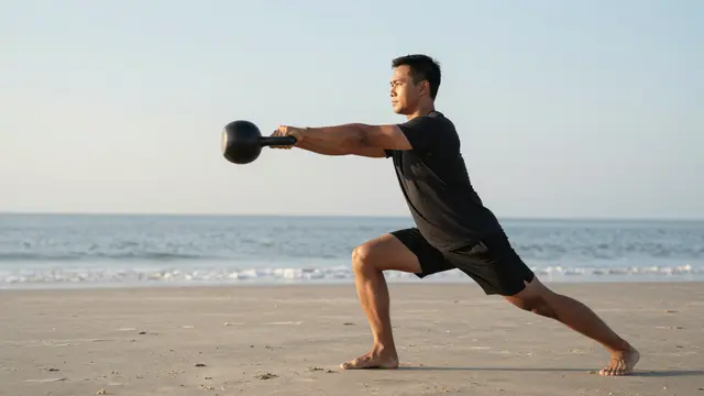 Side view of a Southeast Asian man performing a kettlebell exercise on a sandy beach at sunrise
