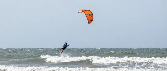 A distant kitesurfer is lifted above breaking waves below a bright orange kite under an overcast sky.