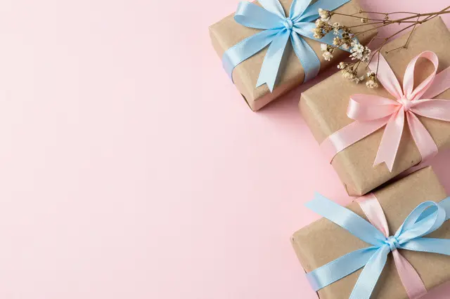 Flat lay of kraft paper wrapped gifts with pastel ribbons and dried flowers on pale pink, with empty copy space on the left.