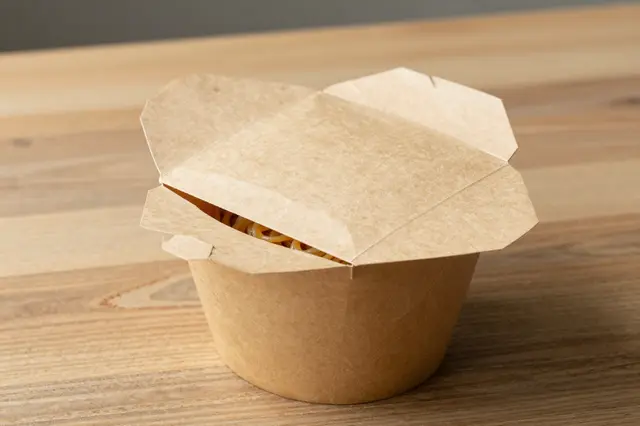 An open kraft paper takeaway box holding noodles sits on a light wooden tabletop.