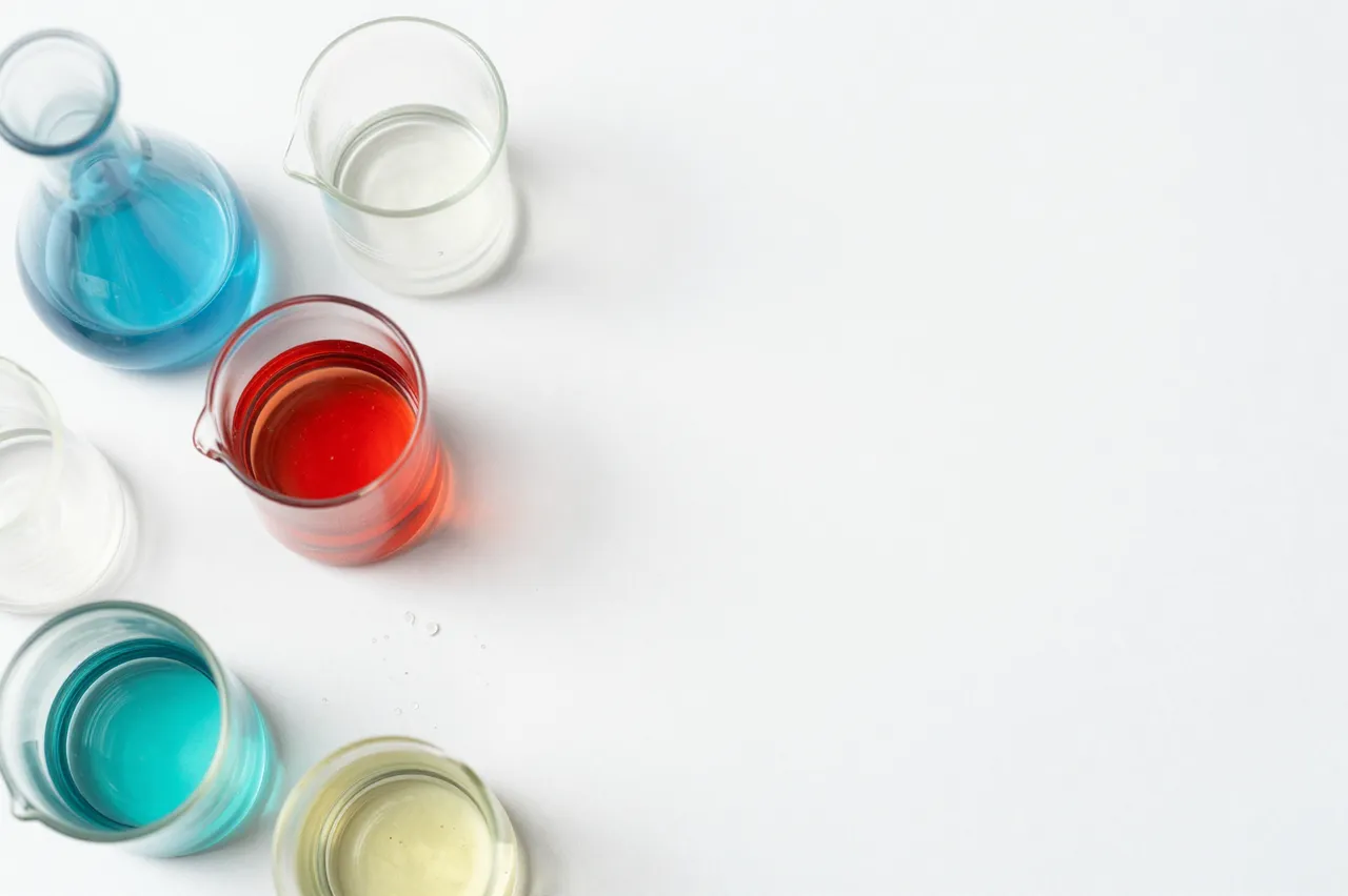 Top down view of unmarked lab beakers and flasks with colored liquids arranged on the left of a plain white surface, leaving