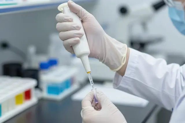 Gloved lab worker uses a pipette to transfer liquid into a small test tube in a laboratory
